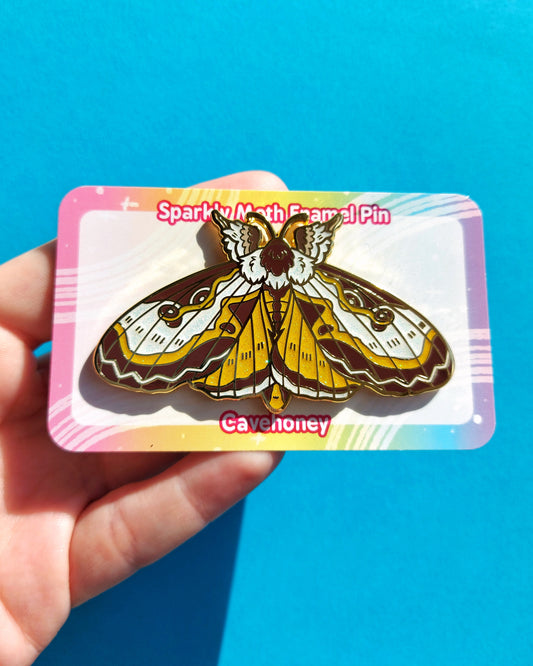 Moth-themed enamel pin in packaging held by a hand against a blue background